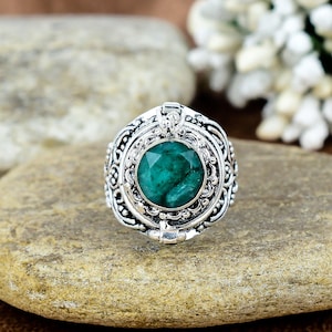 May include: A silver ring with an emerald-cut, deep green gemstone at the center. The ring has an ornate, detailed design around the gemstone and band. The ring is set on a light brown stone surface.