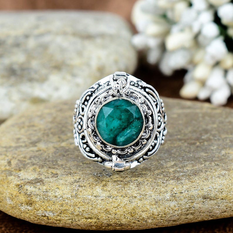 May include: A silver ring with an emerald-cut, deep green gemstone at the center. The ring has an ornate, detailed design around the gemstone and band. The ring is set on a light brown stone surface.