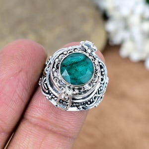 May include: A silver ring with an emerald-cut, vibrant green gemstone. The ring features intricate, ornate detailing around the gemstone and on the band. The ring is a close-up shot, highlighting the craftsmanship and the gemstone's color.