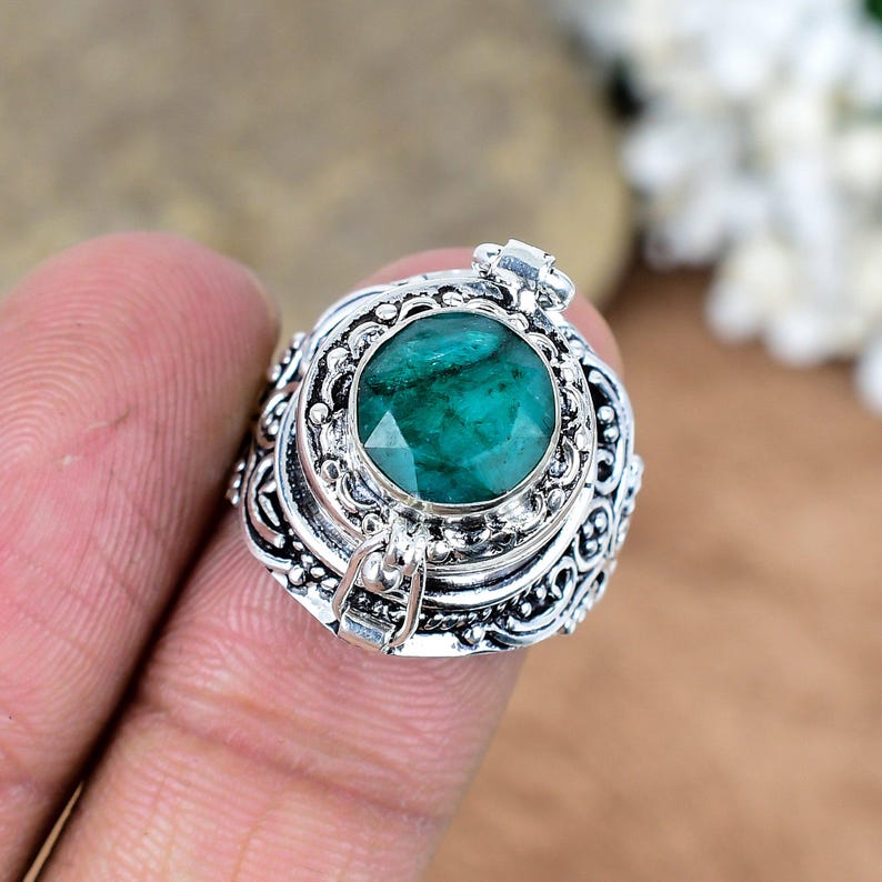 May include: A silver ring with an emerald-cut, vibrant green gemstone. The ring features intricate, ornate detailing around the gemstone and on the band. The ring is a close-up shot, highlighting the craftsmanship and the gemstone's color.