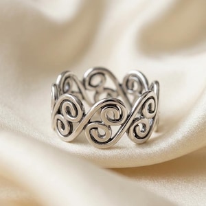 Può includere: Un anello color argento con un design continuo e traforato di motivi a spirale. L'anello è posto su un morbido tessuto bianco sporco, che ne evidenzia i dettagli intricati e la lucentezza metallica. Il design dell'anello è simmetrico ed elegante.