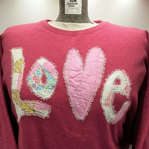 May include: A red sweatshirt with the word "love" appliqued on the front. The letters are made of patchwork fabric in various floral and solid pink patterns. The "o" is a pink heart.