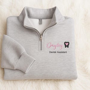 May include: A light gray quarter-zip sweatshirt with the name "Dayley" in pink script, a black tooth with a pink heart, and the words "Dental Assistant" in black. The sweatshirt is folded on a white, fluffy surface.