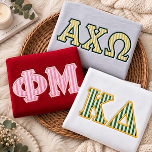 May include: Three folded sweatshirts featuring Greek letters. A light gray sweatshirt with yellow and white striped letters AXΩ, a red sweatshirt with pink and white striped letters ΦM, and a white sweatshirt with green and white striped letters KA.