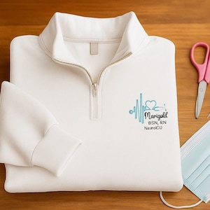 May include: White quarter-zip sweatshirt with a gold zipper and a medical-themed design. The design includes a heartbeat, a stethoscope, and the name "Marigold" with "BSN, RN NeuroICU". A blue pen, pink scissors, a syringe, and a face mask are also visible.