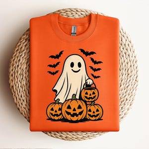 May include: An orange crewneck sweatshirt featuring a graphic of a white ghost holding a small jack-o'-lantern bucket, surrounded by three larger jack-o'-lanterns at the base and several black bats flying in the background above the ghost.