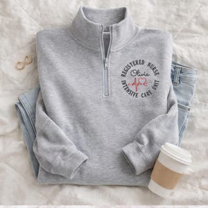 May include: A grey quarter-zip sweatshirt featuring the text "REGISTERED NURSE Olivia INTENSIVE CARE UNIT" in a circular design, with a red heart and heartbeat line. The sweatshirt is shown with blue jeans and a coffee cup.
