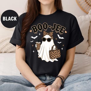 May include: A black t-shirt featuring a graphic of a white ghost wearing leopard print sunglasses, a matching bow, and holding a leopard print handbag and a drink. The text BOO-JEE is printed above the ghost, surrounded by small bats and stars.