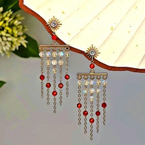 Könnte beinhalten: Ein Paar goldfarbene Chandelier-Ohrringe. Jeder Ohrring hat einen Sonnenstrahl-Stecker, eine rote Perle, einen horizontalen Balken mit klaren und irisierenden Perlen und baumelnde Ketten mit roten Perlen. Der Hintergrund ist neutral.