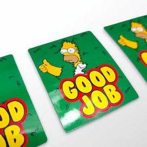 May include: Rectangular green cards featuring Homer Simpson giving a thumbs up, emerging from green foliage. The words "GOOD JOB" are in yellow with a red outline. The cards have a glossy finish.