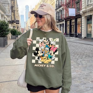 May include: An olive green sweatshirt featuring a checkerboard design with classic Disney characters, including Mickey Mouse, Donald Duck, and Goofy. The words "Mickey & Co." are printed below the characters. The sweatshirt has long sleeves.