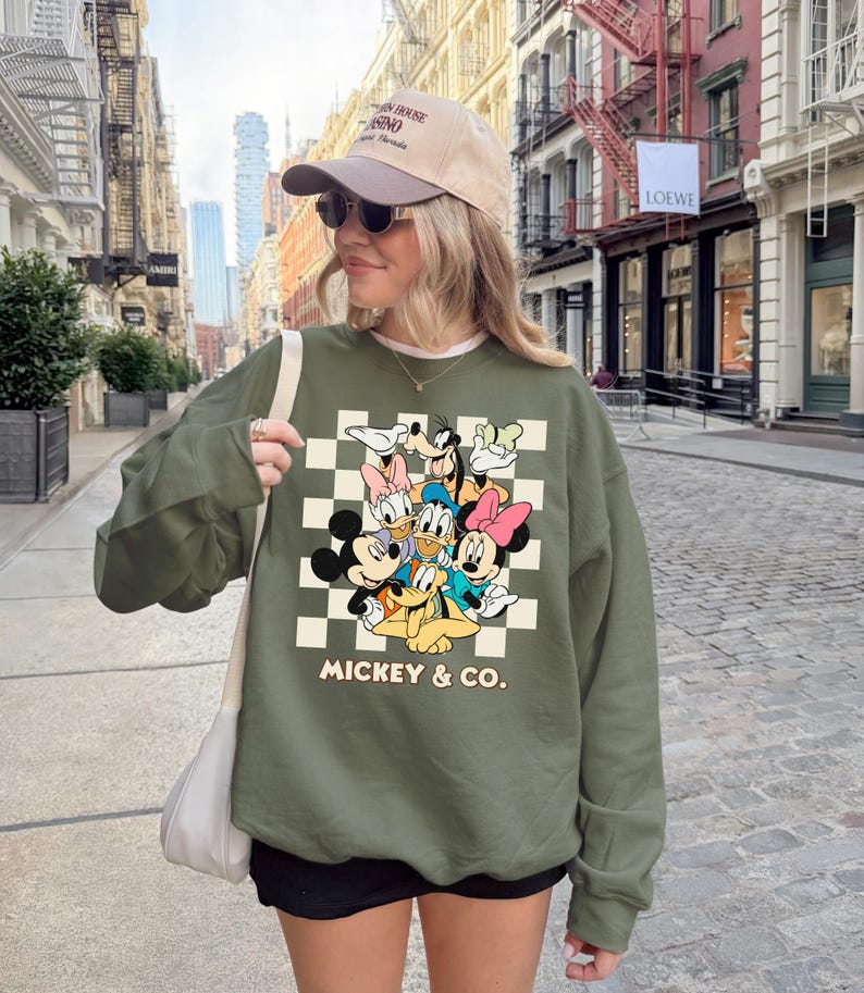 May include: An olive green sweatshirt featuring a checkerboard design with classic Disney characters, including Mickey Mouse, Donald Duck, and Goofy. The words "Mickey & Co." are printed below the characters. The sweatshirt has long sleeves.