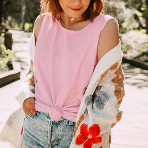 Puede incluir: Una camiseta sin mangas de color rosa claro anudada a la cintura, combinada con pantalones cortos de mezclilla azul con lavado &aacute;cido y un c&aacute;rdigan abierto de color crema con patrones florales texturizados en tonos naranja, azul y marr&oacute;n.