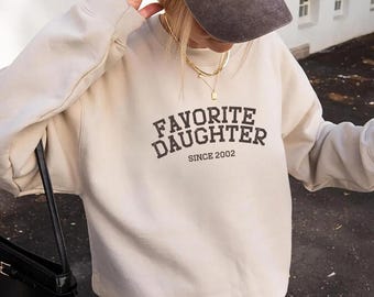 May include: A beige sweatshirt with the text "FAVORITE DAUGHTER SINCE 2002" in dark brown lettering. The image also shows a dark brown baseball cap and a black purse.