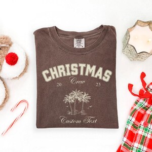 May include: A brown crewneck t-shirt featuring the text CHRISTMAS Crew 20 25 and a graphic of palm trees. The shirt has a Comfort Colors label at the inner neckline. The design is printed in a light cream color on the front of the garment.