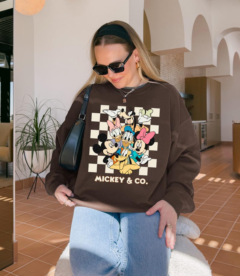 May include: A brown sweatshirt featuring a graphic of Mickey Mouse and friends on a checkered background. The text "MICKEY & CO." is printed below the characters. The sweatshirt is a crew neck style.