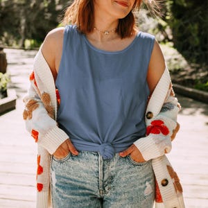 Puede incluir: Una camiseta sin mangas de color azul pizarra s&oacute;lido con un dobladillo anudado, usada debajo de un c&aacute;rdigan abierto de color crema con grandes parches coloridos y texturizados, combinada con jeans de mezclilla azul claro desgastados.