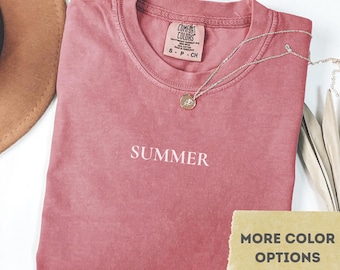 May include: Crimson t-shirt with the word "SUMMER" printed in white. The shirt is folded and has a small tag at the neckline. A silver necklace with a pendant is draped across the shirt. The image also includes a hat and a "More Color Options" tag.