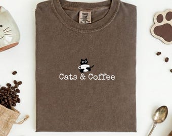 May include: A brown t-shirt with the text "Cats & Coffee" and a graphic of a black cat holding a coffee cup. The shirt is folded and has a tag at the neckline. Coffee beans and a paw-shaped coaster are visible.