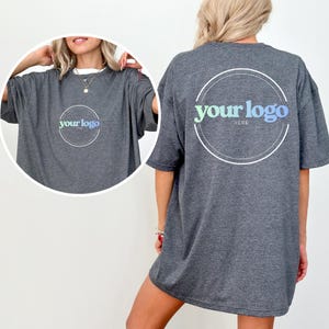 K&ouml;nnte beinhalten: Ein dunkelgraues T-Shirt mit einem wei&szlig;en Rundhalsausschnitt. Auf der Vorderseite befindet sich ein kreisf&ouml;rmiges Logo mit dem Text "your logo" in einem Gr&uuml;n-Blau-Verlauf. Auf der R&uuml;ckseite des T-Shirts befindet sich das gleiche Logo-Design.