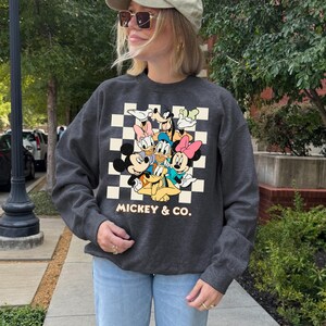 May include: Dark gray crewneck sweatshirt featuring a graphic of Mickey Mouse, Minnie Mouse, Donald Duck, Daisy Duck, Goofy, and Pluto on a checkered background. The text "Mickey & Co." is below the graphic. The sweatshirt is worn by a person.