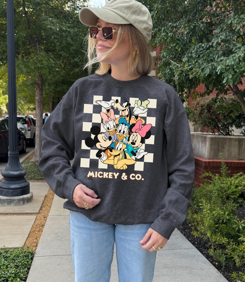 May include: Dark gray crewneck sweatshirt featuring a graphic of Mickey Mouse, Minnie Mouse, Donald Duck, Daisy Duck, Goofy, and Pluto on a checkered background. The text "Mickey & Co." is below the graphic. The sweatshirt is worn by a person.