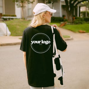 K&ouml;nnte beinhalten: Schwarzes T-Shirt mit einem wei&szlig;en kreisf&ouml;rmigen Logo mit der Aufschrift "your logo here". Die Person tr&auml;gt eine wei&szlig;e Kappe, Sonnenbrille und eine schwarz-wei&szlig;e Tragetasche. Das Bild wurde im Freien aufgenommen.