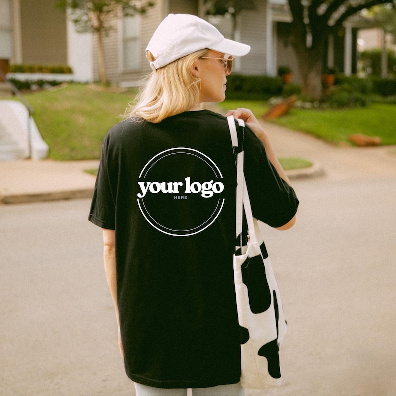 K&ouml;nnte beinhalten: Schwarzes T-Shirt mit einem wei&szlig;en kreisf&ouml;rmigen Logo mit der Aufschrift "your logo here". Die Person tr&auml;gt eine wei&szlig;e Kappe, Sonnenbrille und eine schwarz-wei&szlig;e Tragetasche. Das Bild wurde im Freien aufgenommen.