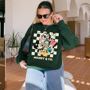 May include: A dark green sweatshirt featuring a graphic of Mickey Mouse and friends on a checkered background. The text "MICKEY & CO." is printed below the characters. The sweatshirt has long sleeves.