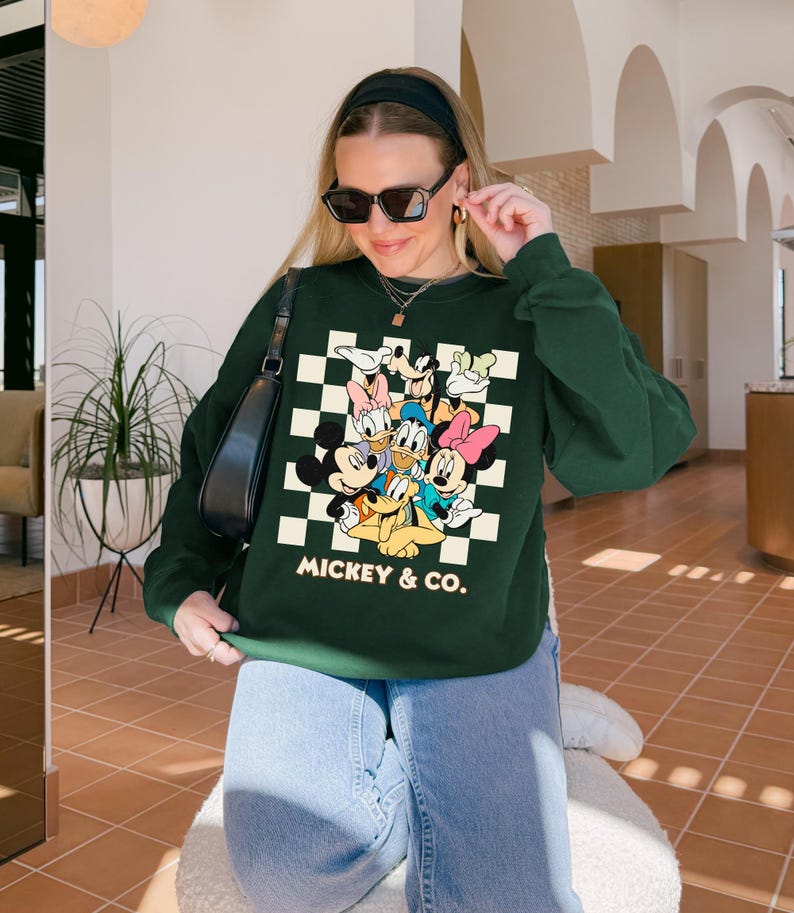 May include: A dark green sweatshirt featuring a graphic of Mickey Mouse and friends on a checkered background. The text "MICKEY & CO." is printed below the characters. The sweatshirt has long sleeves.