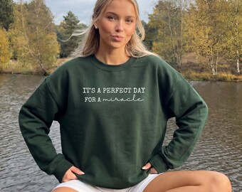 May include: A dark forest green crewneck sweatshirt featuring white text printed on the chest that reads, "IT'S A PERFECT DAY FOR A miracle." The garment has long sleeves, a ribbed neckline, and a ribbed hem, constructed from a solid-colored fabric.