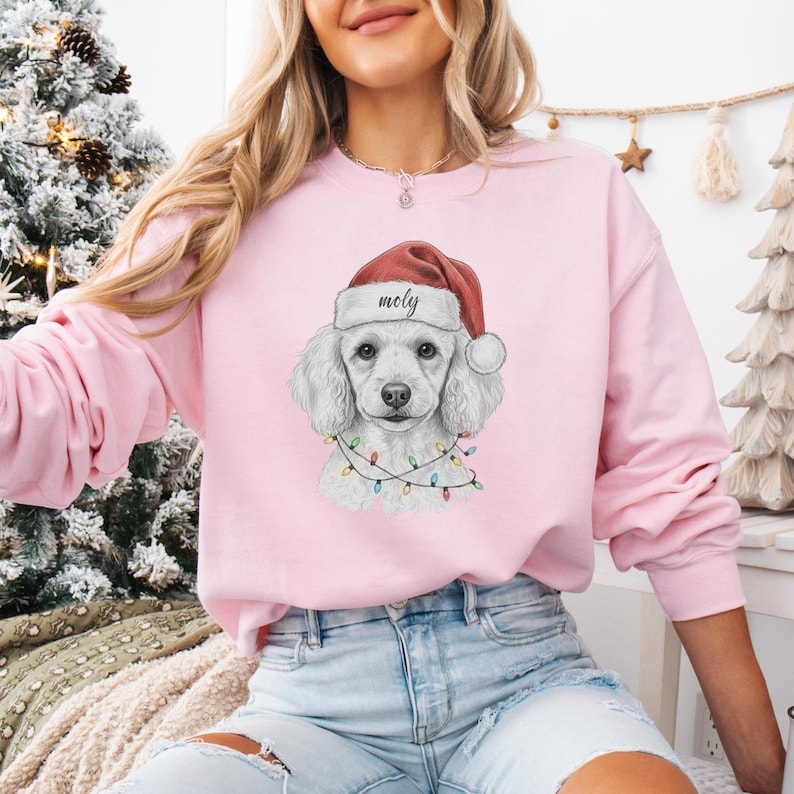 May include: A light pink sweatshirt featuring a white poodle wearing a Santa hat with the name "Moly" and a string of colorful Christmas lights. The poodle is the central graphic on the front of the sweatshirt.