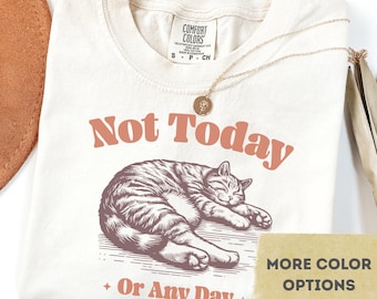 May include: Ivory-colored t-shirt with the text "Not Today" and "Or Any Day" in a retro font. A detailed illustration of a sleeping cat is below the text. A gold necklace with a pendant is draped on the shirt.