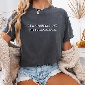 May include: Dark gray t-shirt with the text "IT'S A PERFECT DAY FOR A miracle" in white script. The shirt is a relaxed fit and has short sleeves. The model is wearing denim shorts and a cream-colored cardigan.