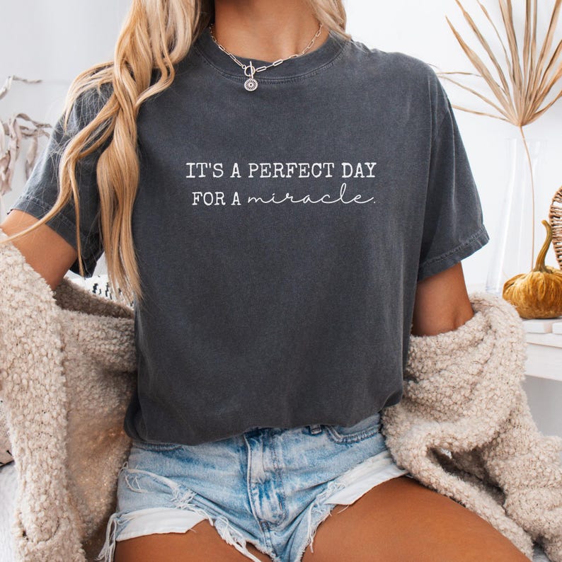 May include: Dark gray t-shirt with the text "IT'S A PERFECT DAY FOR A miracle" in white script. The shirt is a relaxed fit and has short sleeves. The model is wearing denim shorts and a cream-colored cardigan.