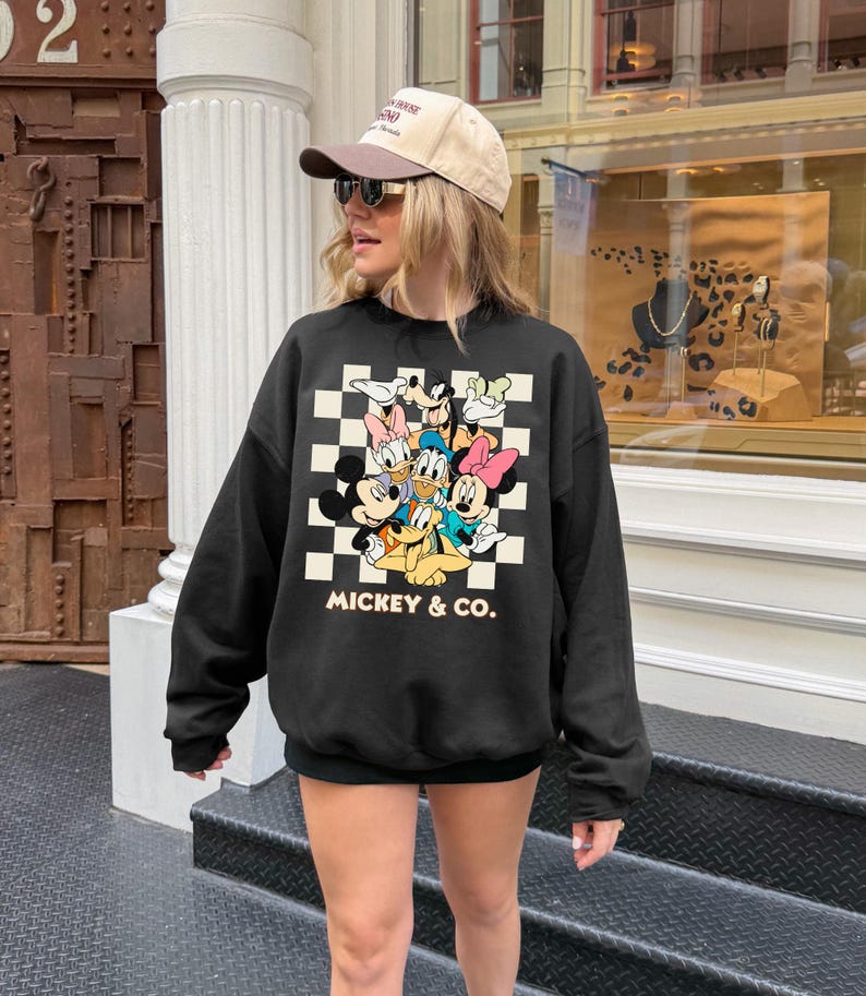 May include: Black sweatshirt featuring a checkered pattern with Mickey Mouse and friends. The sweatshirt has the text "MICKEY & CO." in white. The person in the image is wearing a tan baseball cap and sunglasses.