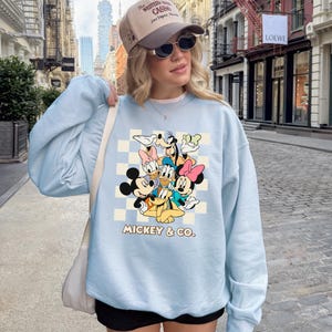 May include: A light blue sweatshirt featuring a graphic of Mickey Mouse and friends. The design includes Mickey, Minnie, Donald, Daisy, Goofy, and Pluto, set against a checkered background. The text "MICKEY & CO." is printed below the characters.