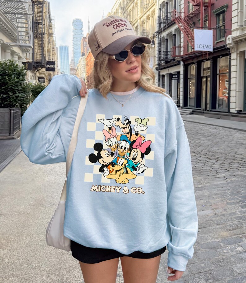 May include: A light blue sweatshirt featuring a graphic of Mickey Mouse and friends. The design includes Mickey, Minnie, Donald, Daisy, Goofy, and Pluto, set against a checkered background. The text "MICKEY & CO." is printed below the characters.