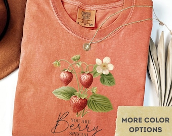 May include: A yam-colored Comfort Colors t-shirt featuring a strawberry plant illustration with the words "You are Berry Special." A gold necklace and a tag that says "More Color Options" are also visible.