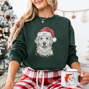 May include: Dark green sweatshirt featuring a Golden Retriever wearing a Santa hat with the name "Ares" and a string of Christmas lights. The dog's portrait is centered on the sweatshirt. The sweatshirt is paired with red and white plaid pajama pants.