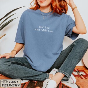 May include: A blue t-shirt with the text "don't hear what I didn't say" in white. The shirt is a solid color and has short sleeves. The person in the image is wearing blue jeans and white sneakers.