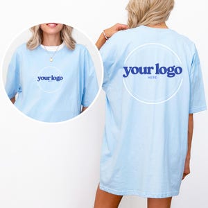 K&ouml;nnte beinhalten: Hellblaues T-Shirt mit dem Text "your logo" in einem wei&szlig;en Kreis auf der Vorder- und R&uuml;ckseite. Das Shirt ist einfarbig und hat kurze &Auml;rmel. Der Text "HERE" befindet sich unter dem Logo auf der R&uuml;ckseite.