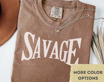 May include: Espresso-colored t-shirt with the word "SAVAGE" in large, white, block letters. The shirt has a crew neck and short sleeves. A silver necklace with a small pendant is draped across the shirt. The shirt is labeled "Comfort Colors".