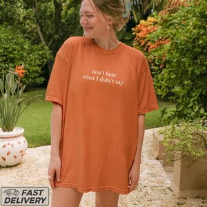May include: An orange t-shirt with the text "don't hear what I didn't say" in white. The shirt is a solid color and has short sleeves. The image is taken outdoors with greenery in the background.