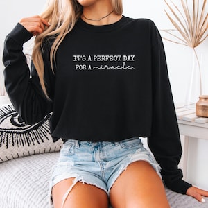 May include: Black long-sleeve sweatshirt with the text "IT'S A PERFECT DAY FOR A miracle" in white script. The sweatshirt is paired with denim shorts. The image is of a person wearing the sweatshirt.