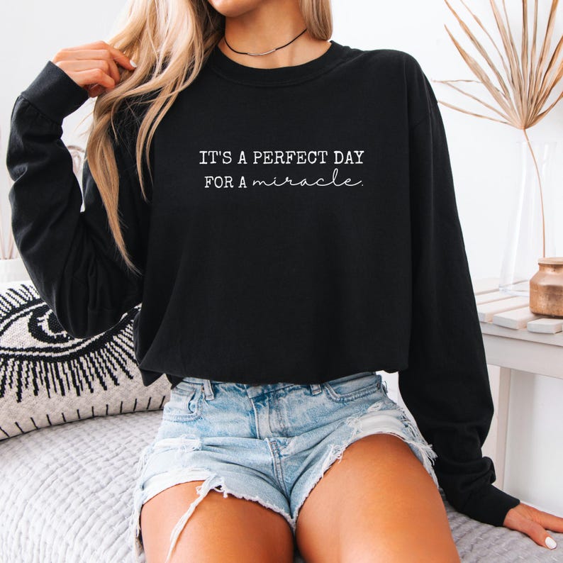 May include: Black long-sleeve sweatshirt with the text "IT'S A PERFECT DAY FOR A miracle" in white script. The sweatshirt is paired with denim shorts. The image is of a person wearing the sweatshirt.