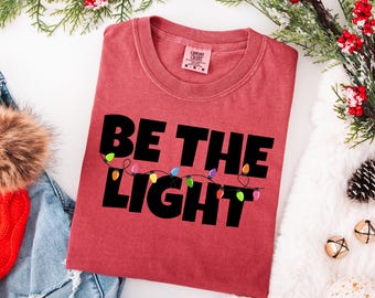 Peut inclure: Un t-shirt rouge avec l'inscription "BE THE LIGHT" en grandes lettres noires. Le mot "LIGHT" est orné d'une guirlande de lumières de Noël colorées. Le t-shirt est entouré de décorations de Noël.