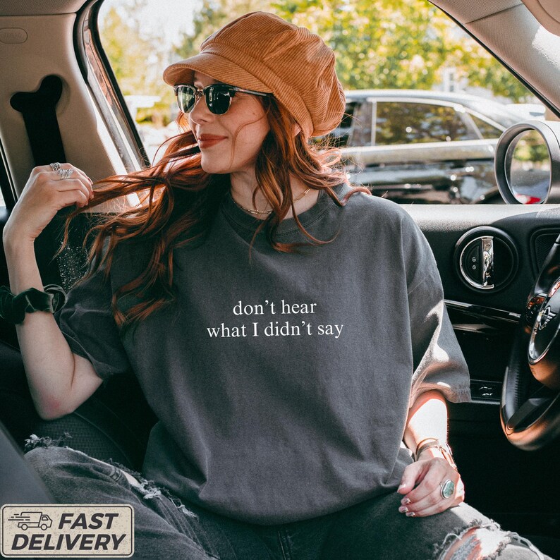 May include: Dark gray t-shirt with the text "don't hear what I didn't say" in white. The person in the image is wearing sunglasses and a brown corduroy hat. The person is sitting in a car.