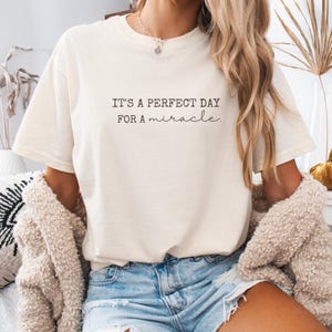 May include: Cream-colored t-shirt with the text "IT'S A PERFECT DAY FOR a miracle" in black. The shirt has short sleeves and a relaxed fit. The model is wearing a necklace and a cream-colored cardigan.