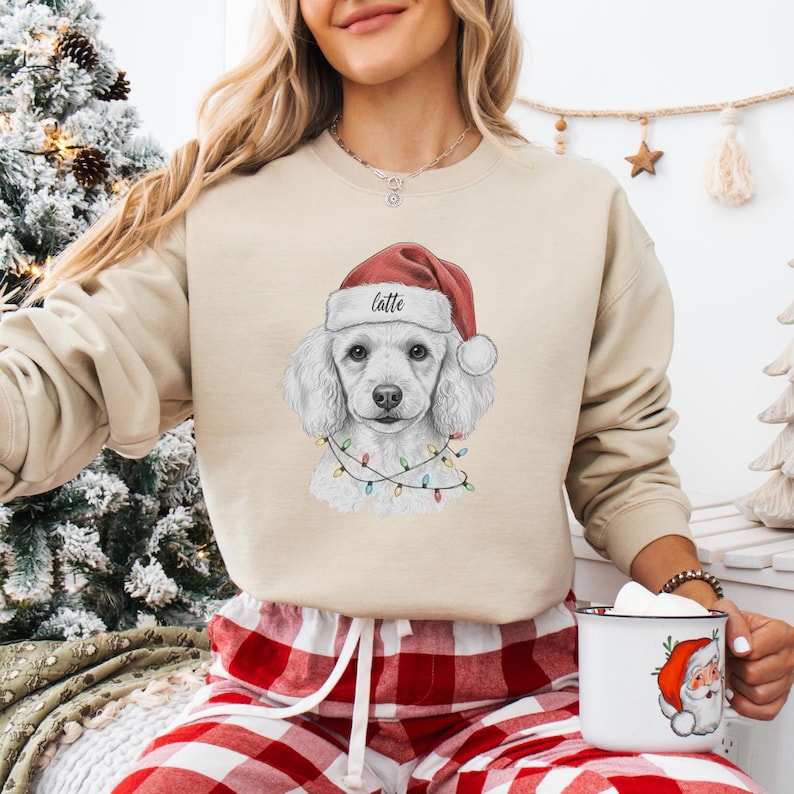 May include: Beige sweatshirt featuring a white poodle wearing a Santa hat with the word "latte" and a string of Christmas lights. The person is wearing red and white plaid pajama pants and holding a mug with a Santa Claus design.
